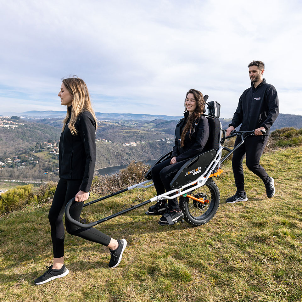 A Joëlette Adventure mono-wheel all-terrain wheelchair being operated by two guides on a grassy hillside. One guide pulls from the front using long handlebars, while the other pushes and balances from the rear. The rider is seated in a central chair directly above the single off-road tire, demonstrating the specialized teamwork required for technical single-track hiking.