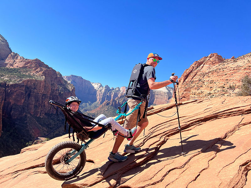 A Huckleberry Hiker mono-wheel hiking chair being pulled up a steep red rock slope in Zion National Park. A man wears a specialized trekking harness to pull the chair while a young girl sits comfortably in the seat over a single large fat-tire wheel. The iconic sandstone cliffs of Zion are visible in the background under a clear blue sky.