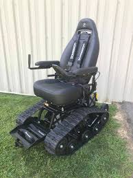 A TracFab electric tracked wheelchair positioned on asphalt against a vibrant orange and purple sunset. The chair features heavy-duty black tank treads, a black racing-style bucket seat with blue four-point safety harnesses, and a joystick controller, highlighting its rugged build and independent power for all-terrain use.