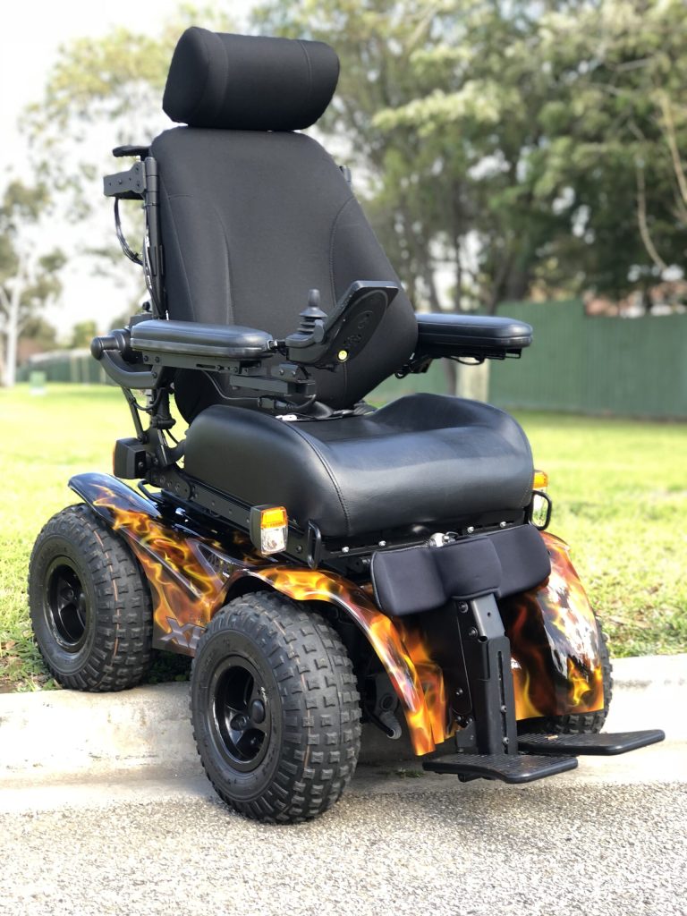 A custom Magic Mobility Extreme X8 all-terrain power wheelchair with a "fire and flames" hydrographic wrap on the fenders. The chair is shown demonstrating its 4x4 curb-climbing ability with its front left knobby tire perched on a concrete curb while the other wheels remain on the asphalt, highlighting its articulating front frame.