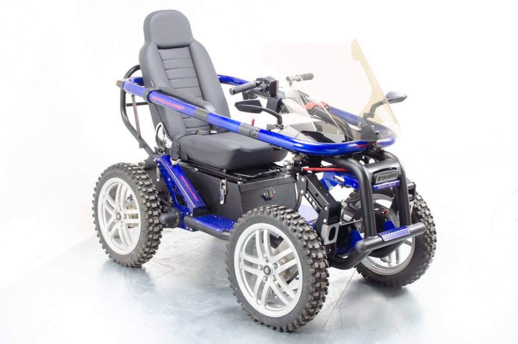 A blue and black TerrainHopper Overlander 4ZS 4x4 off-road wheelchair in a studio setting. The vehicle features four independent knobby off-road tires, a protective blue roll-bar frame, a clear windshield, and an ATV-style handlebar steering system, designed for extreme outdoor independence.