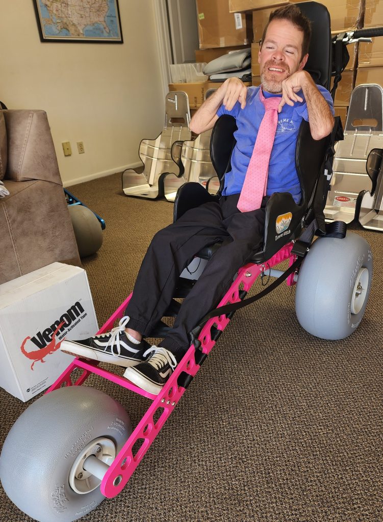 Man with cerebral palsy sits in Ainsley's Angels wheelchair. He's wearing a pink tie to match the pink wheelchair.