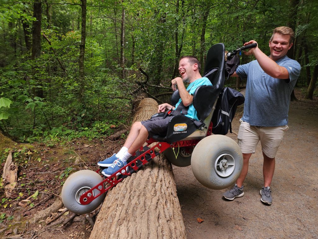 A man with cerebral palsy sits in a Extreme Motus off-road wheelchair. He is  being pushed over a large log in the Great Smoky Mountain National Park. 