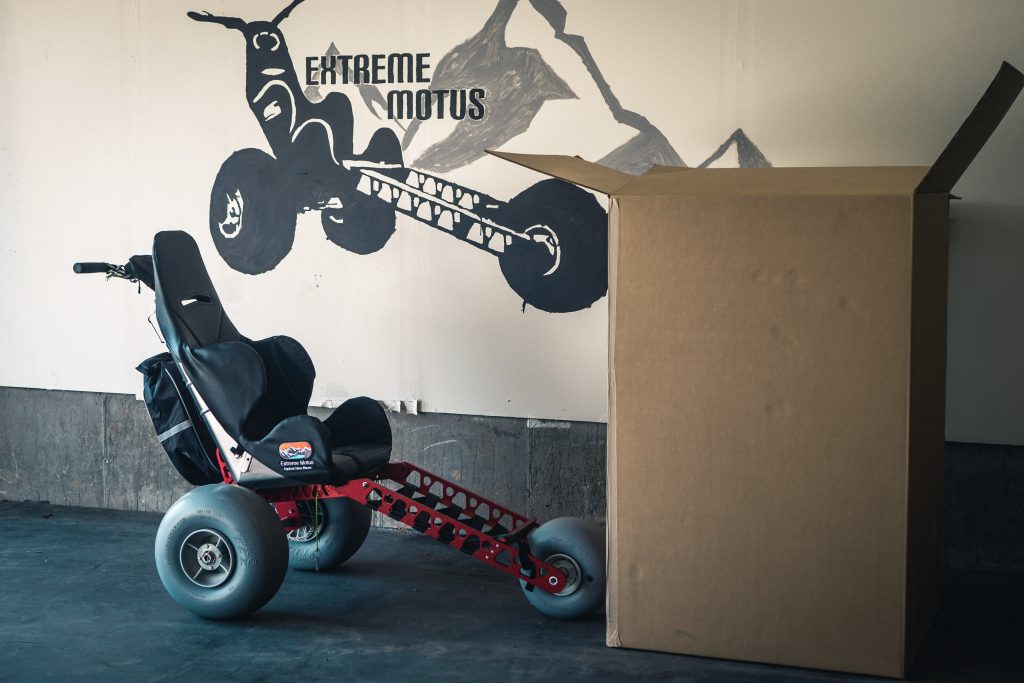 The Extreme Motus all terrain wheelchair sitting next to a large cardboard box. Painted on the wall behind the wheelchair and box is the Extreme Motus logo. 