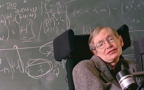 Stephen Hawking sitting in a wheelchair in front of a blackboard with chalk writing on it. 