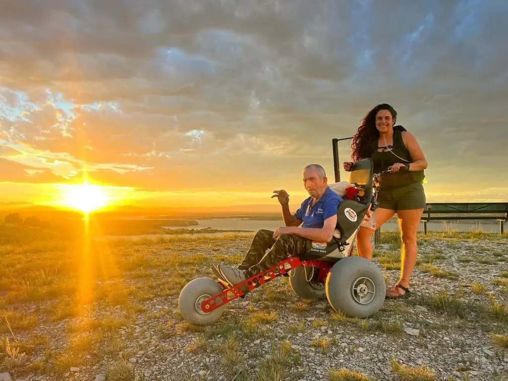 A man in an all-terrain wheelchair and a smiling woman stand on a grassy hilltop at sunset. The sun is low on the horizon, casting a bright golden glow over a valley with a lake in the distance.