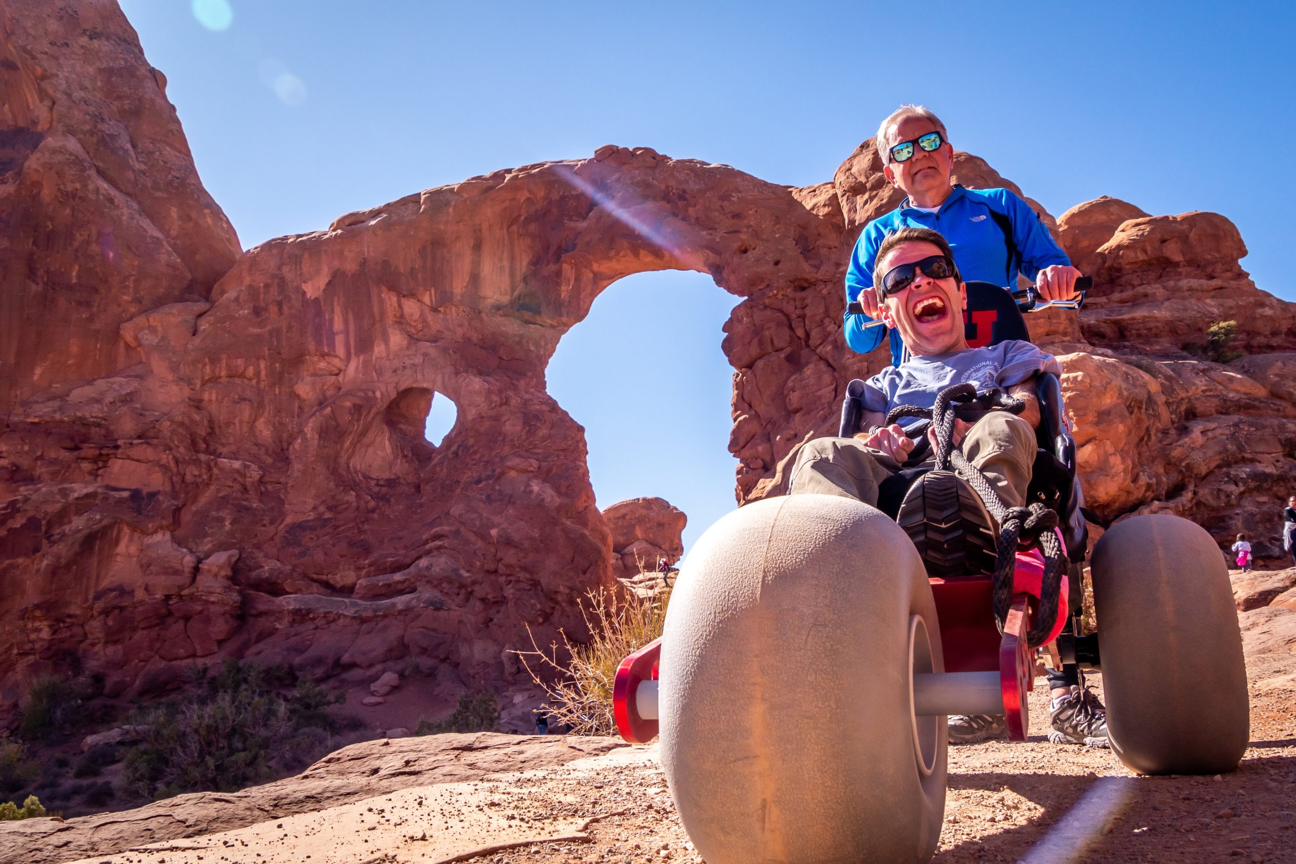 man laughing with joy in a red Extreme Motus all-terrain wheelchair at Arches National Park in Moab, Utah, as his father pushes him toward a large sandstone arch.
