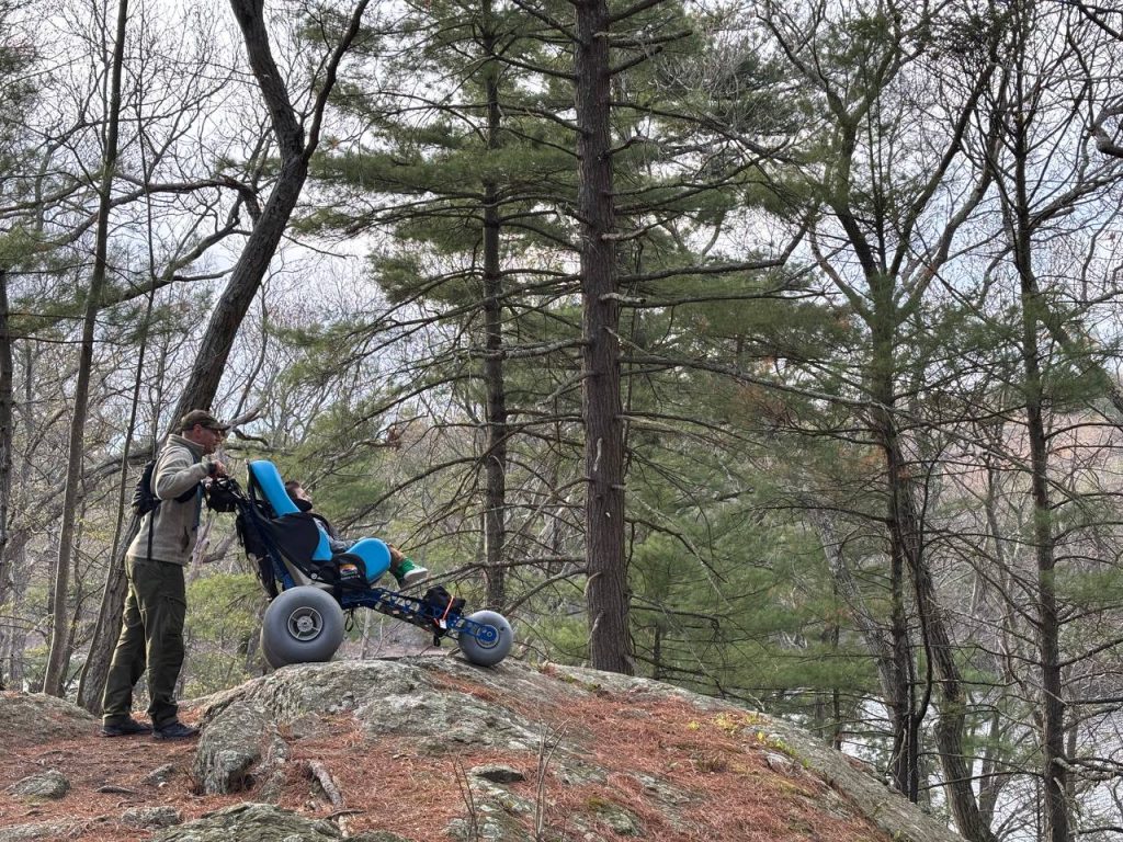 A man in outdoor gear pushes a specialized all-terrain wheelchair with large, balloon-style tires over a rocky, pine-needle-covered ridge. A child sits in the bright blue adaptive seat, looking out over a wooded valley.