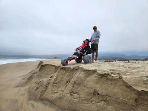Addisyn's father standing beside her on a foggy beach dune in her Extreme Motus all-terrain wheelchair