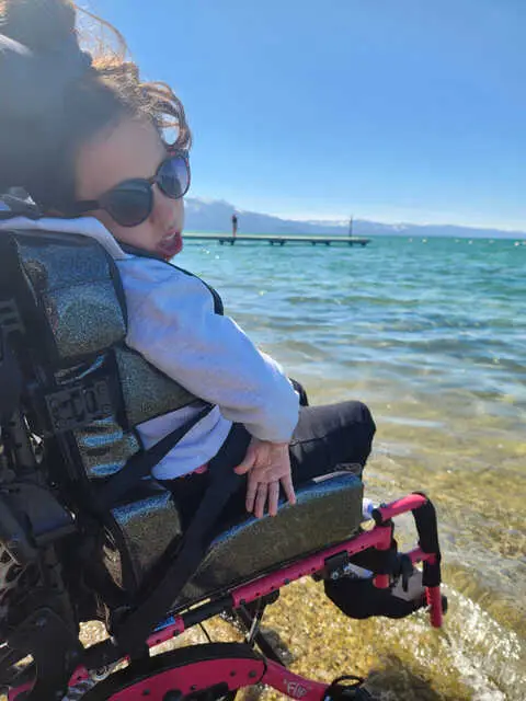 Addisyn smiling in the sun beside a mountain lake in her Extreme Motus all-terrain wheelchair