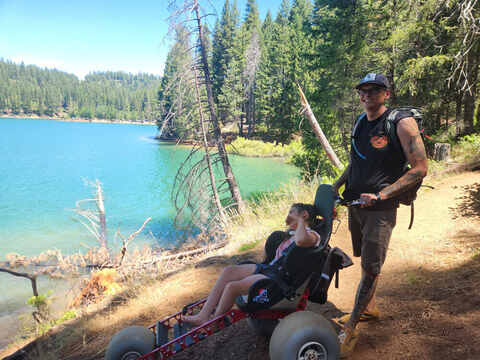 Addisyn's father pushing her along a scenic turquoise lake trail in her Extreme Motus all-terrain wheelchair