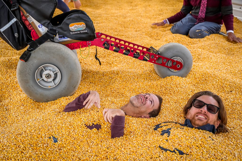 Sam and Ryan buried up to their necks in the Cornbelly's corn pit next to an Extreme Motus all-terrain wheelchair